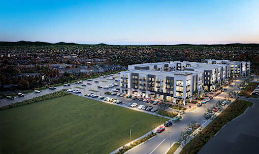A modern building complex is lit up at dusk with cars parked in the lot.
