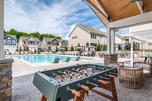 A foosball table is in the middle of a patio with a pool and houses in the background.