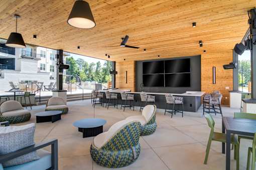 Sugar Hill, GA, Pet-Friendly Apartments - The Collier Sugar Hill - Spacious Outdoor Lounge with Wooden Ceiling, Recessed Lighting, Pendant Lamps, Ceiling Fan, Armchairs, Lounge Chairs, Low Coffee Tables, Square Dining Tables and Chairs, Speaker, Wall-Mounted Lighting, and a Bar Area with Wall-Mounted Screens, White Countertop, and Multiple Chairs.