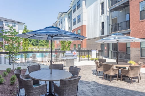 Apartments For Rent In Hermitage, TN - Outdoor Patio Area By The Pool. Patio Has Gas BBQ Grills, Tables With Umbrellas, Chairs, And Maintained Landscaping.