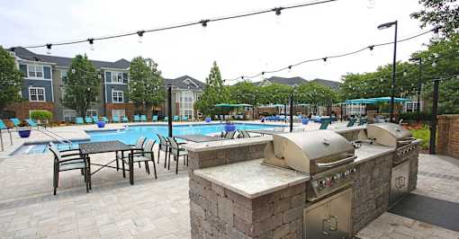 A pool with a barbecue grill and chairs is surrounded by apartment buildings.