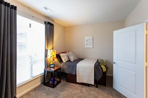 Dog-Friendly Apartments in Johnson City, TN - University Edge - Private Bedroom With Made Bed, Accent Pillows, Soft Carpet, Side Table Lamp, And Large Window With Blinds And Dark Curtains.