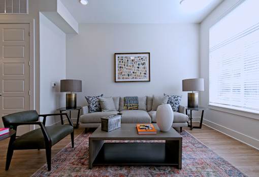 One Bedroom Apartments in Memphis, TN - The Rise - Living Room with Plush Gray Couch, Two Round Side Tables with Lamps, Wooden Coffee Table, Wide Leather-Style Chair, Hardwood-Style Flooring, Area Rug, and Large Window with White Blinds.