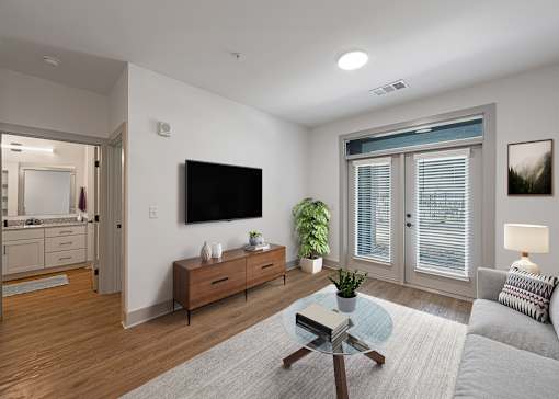 Two-Bedroom Luxury Apartments in Huntsville, AL - The Kelvin - Living Room with Luxury Vinyl Tile Flooring, Area Rug, Sofa, Glass-Top Coffee Table, Side Table with Lamp, Media Cabinet, Wall-Mounted TV, and French Doors with Access to Patio.