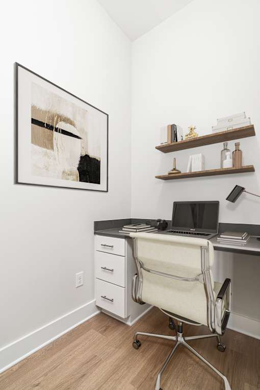 A black and white abstract painting hangs on a white wall above a desk with a laptop and a white chair.