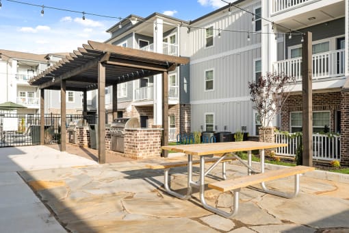Three-Bedroom Luxury Apartments in Alcoa, TN - Vintage Hunters Crossing - Outdoor Patio with Grilling Stations, Pergola, Picnic Table, String Lights, Metal Fence, and View of the Apartments.
