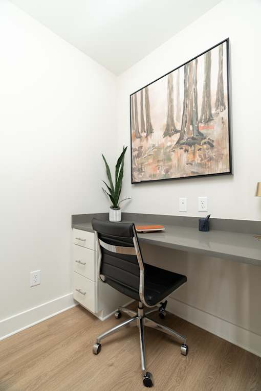A painting of trees hangs on the wall above a desk with a chair and a plant.