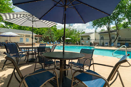 A table with chairs and an umbrella is in the foreground of a pool area.