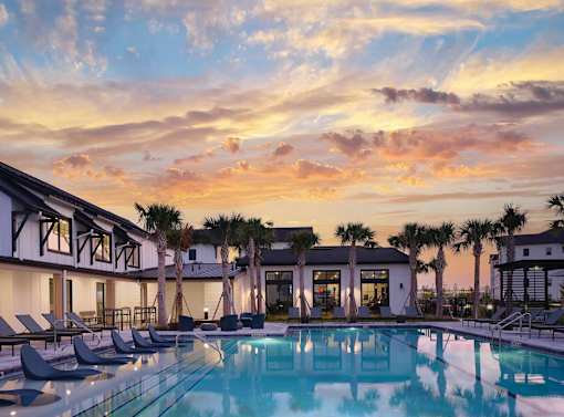 A pool surrounded by lounge chairs and palm trees under a sunset sky.