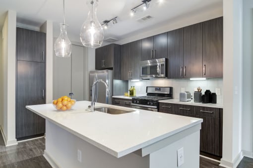 Luxury Apartments Near Piedmont Park In Atlanta, GA - Venue Brookwood - Kitchen With Wood-Grain Flooring, Designer Finishes, Wood-Tone Flat-Panel Custom Cabinetry, Quartz Countertops, Stainless Steel Whirlpool Appliances, Built-in Microwaves, Designer Tile Backsplash With Under-Cabinet Lighting, Chrome Track Lighting, And Gourmet Kitchen Island with Pendant Lighting.