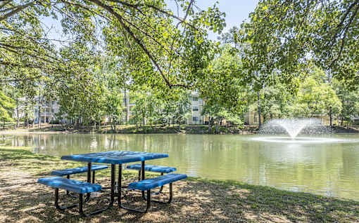 A blue picnic table is set up by a pond with a fountain in the background.