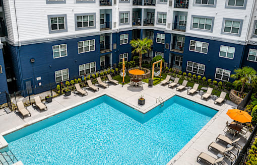 A large swimming pool surrounded by lounge chairs and umbrellas in front of a multi-story building.
