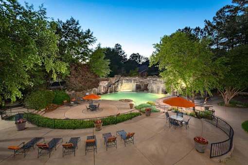 Columbus Apartments - Exterior View Of The Pool Area. The Area Has Lounge Chairs, Patio Sets With Umbrellas, Potted Plants, A Waterfall, Stairs, And Trees.