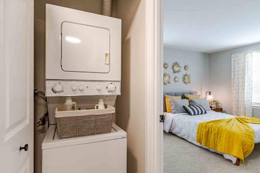 A small white washing machine is in a cabinet in a bedroom with a bed and a window.