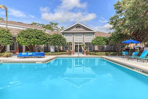 A swimming pool in front of a building with a blue umbrella and chairs.