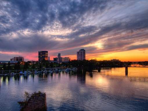 A city skyline at sunset with buildings reflecting in the water.