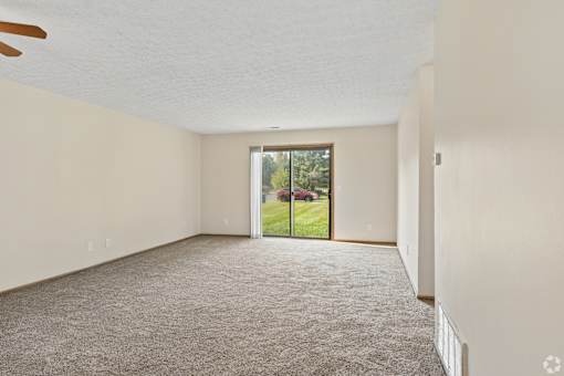 A spacious living room with a carpeted floor, ceiling fan, and a sliding glass door leading to a backyard.