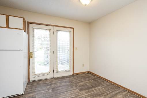 A white refrigerator is in the corner of a room with wood flooring and white walls.