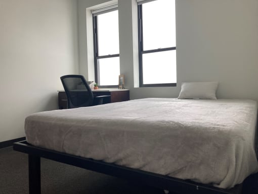 Full extra long bed that is 54 by 80 inches is to right. Covered in gray blanket with white pillow at top. Brown desk with white top to left with desk chair underneath. Two windows are set above desk.