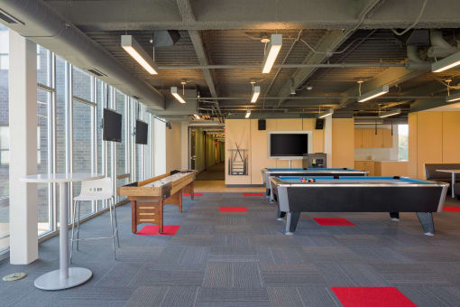 On left, Shuffleboard and high-rise table, white. On right, two pool tables with wall-mounted tv in background. Floor is primarily gray with red squares