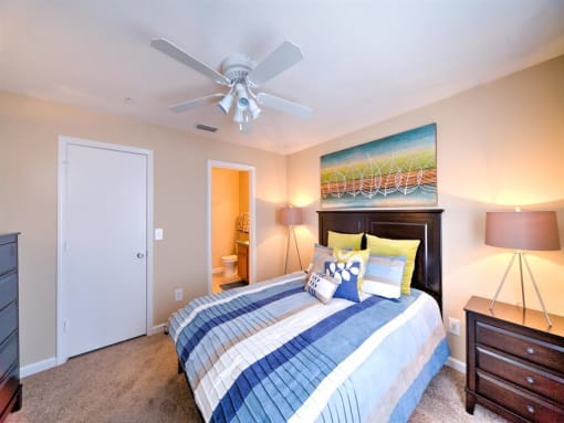 Furnished model bedroom with large closet, in-suite bathroom, carpet flooring, multi speed ceiling fan, side tables, desk lamps, and wall art