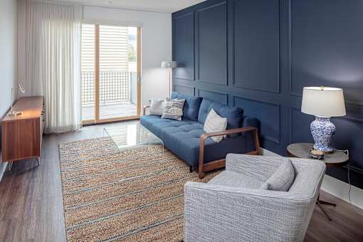 A living room with a grey sofa, a grey armchair, a wooden side table, and a large window with curtains at The Fitz Apartments, Portland