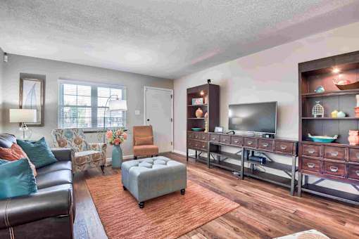 A living room with a brown leather couch, a grey ottoman, a wooden floor, and a television on a stand.
