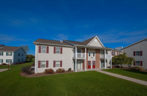 Two-story apartment building with vinyl siding and brick accents, featuring shutters, patios, and balconies. The landscaping is neat, with modern design elements throughout.