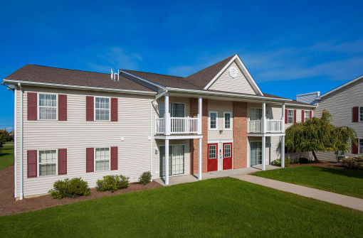 Two-story apartment building with vinyl siding and brick accents, featuring shutters, patios, and balconies. The landscaping is neat, with modern design elements throughout.