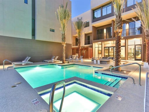 Scottsdale, AZ, Apartments for Rent - Dusk Scottsdale - Dusk View of Resort-Inspired Pool with Spa, White Lounge Chairs, Palm Trees, and Apartment Building in the Background.