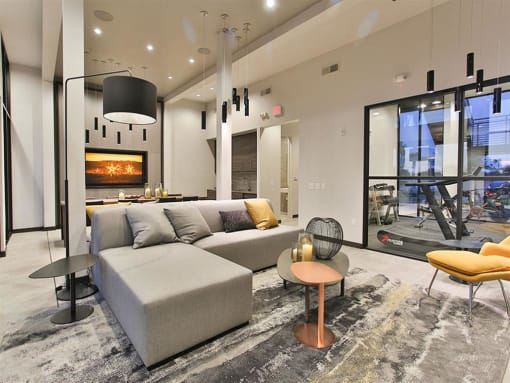 Apartments for Rent in Scottsdale, AZ - Dusk Scottsdale - Resident Lounge with Marbled Area Rug, Grey Sectional Couch, Coffee Table, Standing Lamp, Yellow Cushioned Chair, Table with Chairs, Mounted Wall Art, and View of Gym.