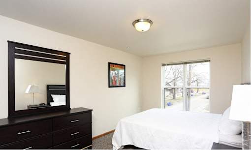 A bedroom with a white bed, a black dresser with a mirror, and a window with a view of trees.