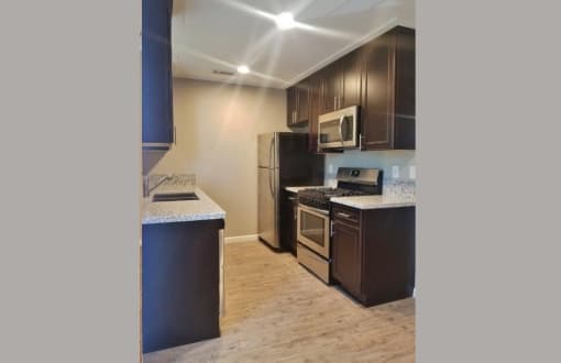Angled view of kitchen area with brown cabinets, stone counters, stainless appliances and wood look flooring