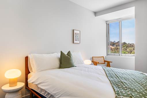 A bedroom with a large bed, a nightstand with a lamp, a picture on the wall, and a window with a view of the outdoors.