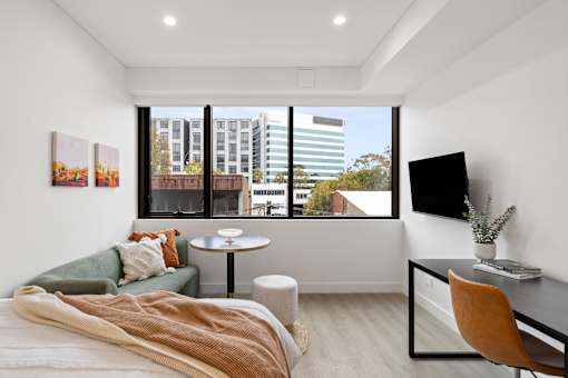 A bedroom with a bed, desk, and chair with a window overlooking a cityscape.