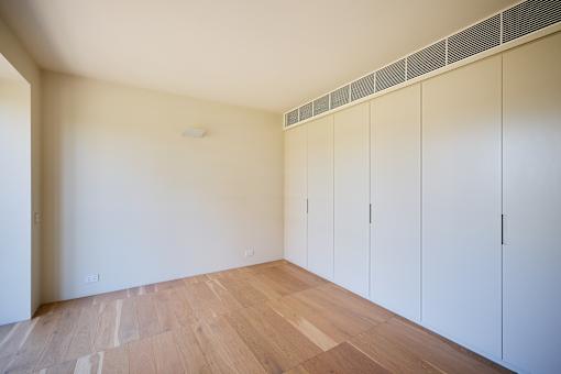 A room with white walls and wooden flooring with a row of white cupboards.