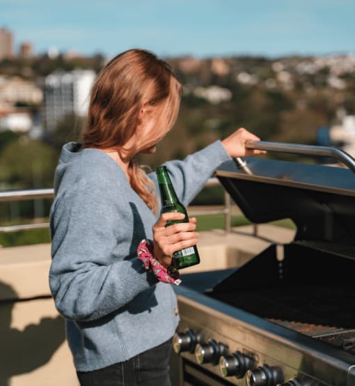 a woman holding a bottle of beer standing next to a grill