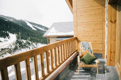A chair with a green pillow is on a balcony overlooking a snowy mountain.