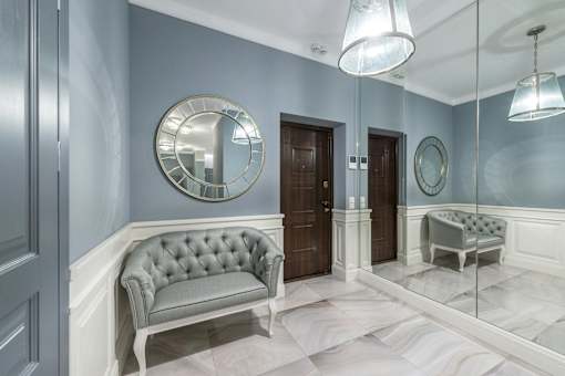 A grey tufted sofa sits in a hallway with a mirrored wall and a round mirror above it.