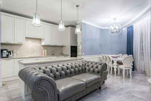 A grey tufted sofa sits in a kitchen with white cabinets and a marble floor.