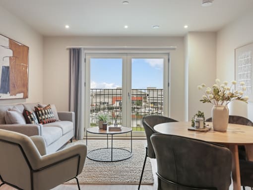 A living room with a grey couch, a round table with a vase of flowers on it, and a balcony with a view of the city.