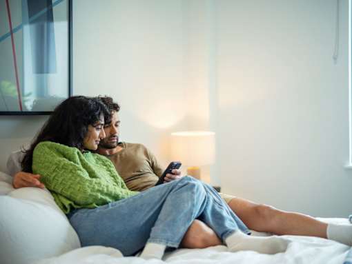 A man and woman are sitting on a bed, with the woman holding a remote control.