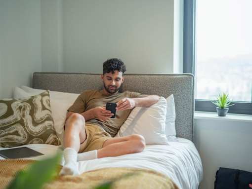 A man is sitting on a bed with a laptop and a phone in his hands.