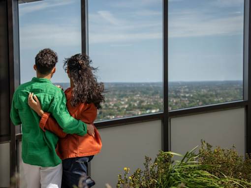 A couple in a green and orange jacket are standing close together and looking out a window at a cityscape.