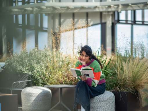 A woman is sitting on a grey stool reading a book in a sunny outdoor area.