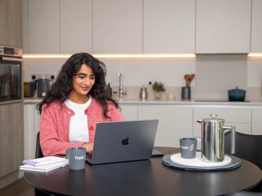 A woman in a pink cardigan is working on her laptop at a table with a cup of coffee and a tray of coffee cups.