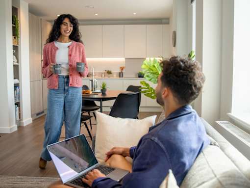 A woman standing in a kitchen holding a mug while a man sits on a couch using a laptop.