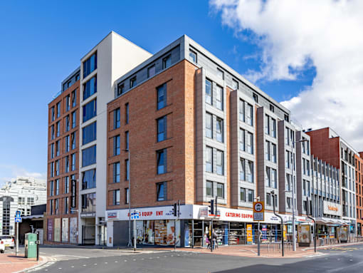 A modern multi-story building with a mix of brick and white panels is situated on a street corner with shops in the foreground.