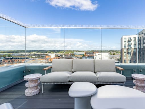 A white couch with a wooden table and two white stools are on a balcony with a glass railing.