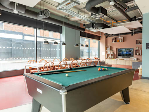 A pool table with a green cloth and snooker balls is in the foreground of a room with a brick wall and large windows.